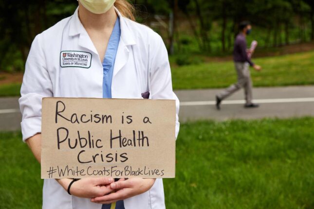 A medical student wearing a white coat with the Washington University School of Medicine emblem holds a sign that states "Racism is a public health crisis. #WhiteCoatsForBlackLives"