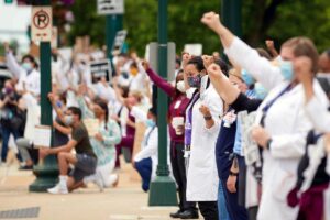 Hundreds of people, some wearing scrubs and white coats, stand on a sidwalk with signs and raised fists.